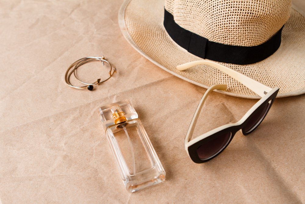 یک بطری عطر شیشه‌ای با درب طلایی، کنار یک جفت عینک آفتابی مشکی با دسته‌های سفید، یک کلاه حصیری با نوار مشکی و چند دستبند نقره‌ای و طلایی روی یک سطح کاغذی بژ قرار گرفته‌اند. این چیدمان حس و حال تابستانی را منتقل می‌کند."
