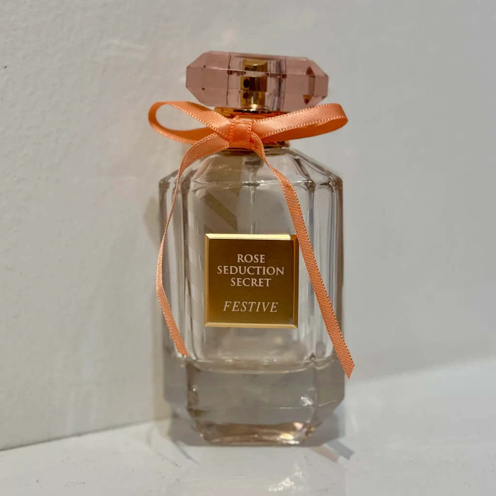 بطری 100 میل عطر زنانه فراگرنس ورد رز سداکشن سکرت فستیو مشابه ویکتوریا سکرت بامبشل سلبریشن با طراحی شیک و صورتی در پس‌زمینه‌ای مینیمال.